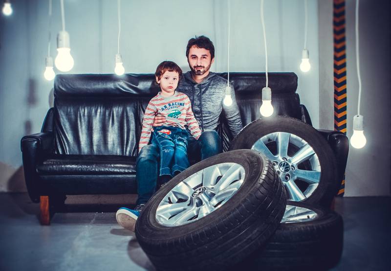 Un hombre adunto con un niñi en sus brazos sentados en un sillón. Grente a ellas hay una pila de neumáticos.