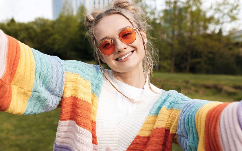 Una mujer joven, rubia, con lentes y un chaleco de colores sostiene la cámara y mira con una gran sonrisa al lente. Está de día en medio de un paisaje exterior vegetado.
