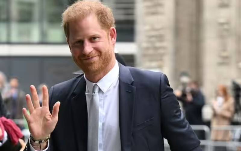 Príncipe Harry en exterior, saludando con un gesto de mano y el rostro sontiente.