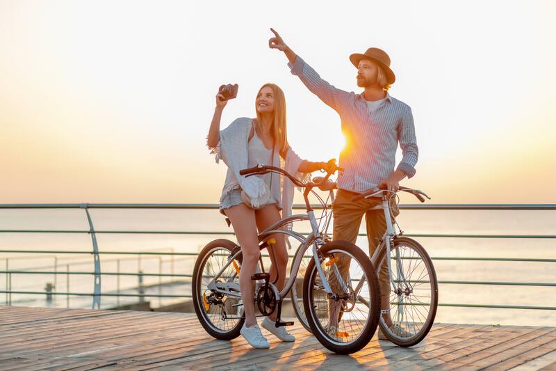 attractive happy couple traveling in summer on bicycles, man and woman with blond hair boho hipster style fashion having fun together, taking photos sightseeing