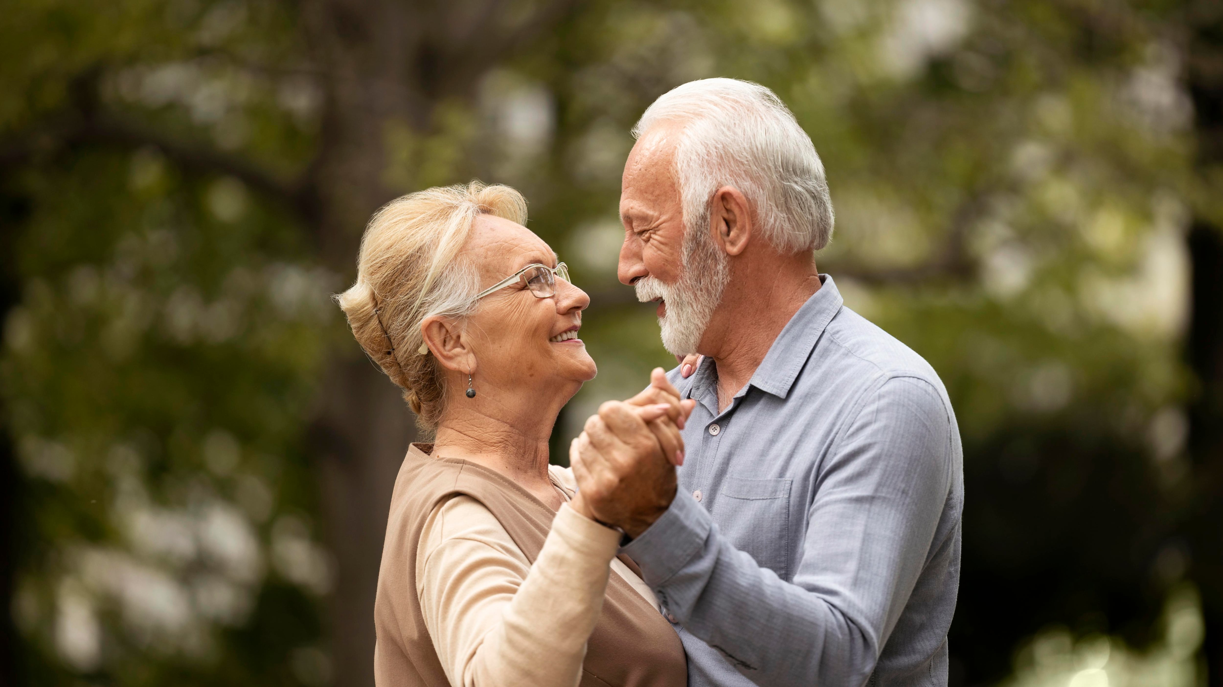 Una pareja de adultos mayores está en un paisaje externo. Tienen sus manos tomadas a la altura de los hombros c omo si estuvieran bailando y se miran a los ojos con amor.