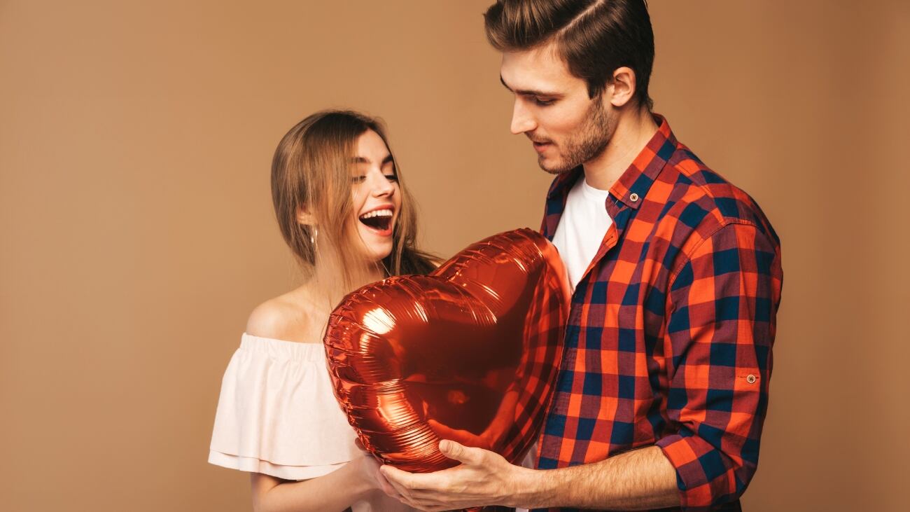 Pareja héterosexual. Ella recibe de él un globo en forma de corazón con cara de alegre sorpresa.