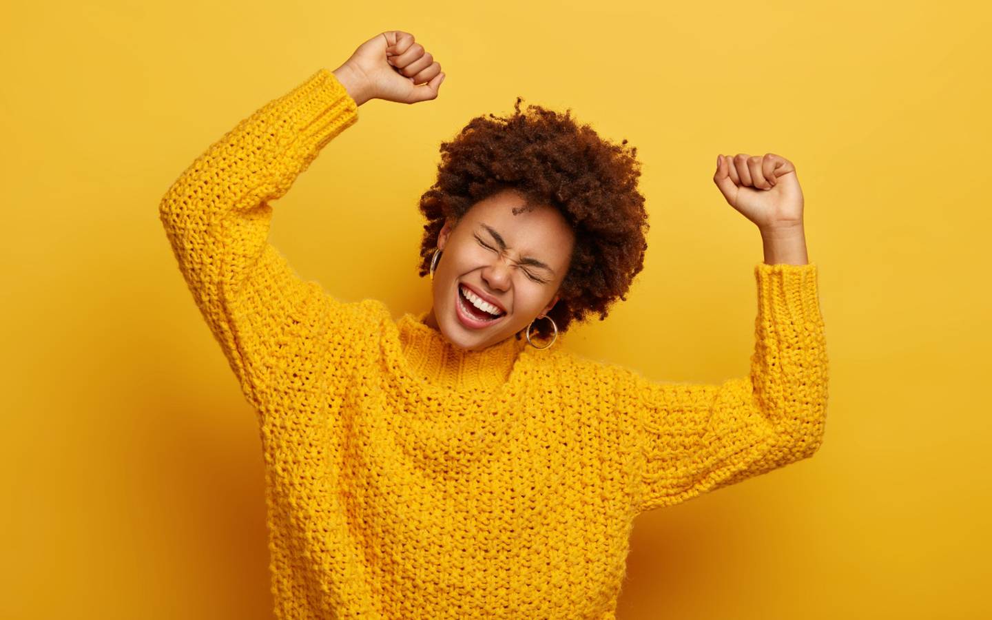 Mujer afrodescendiente de pelo rizado y corto con las manos levantadas en señal de trunfo y con una gran sonrisa de alegría en el rostro. Lleva un sweter amarillo sobre un fondo del mismo color.